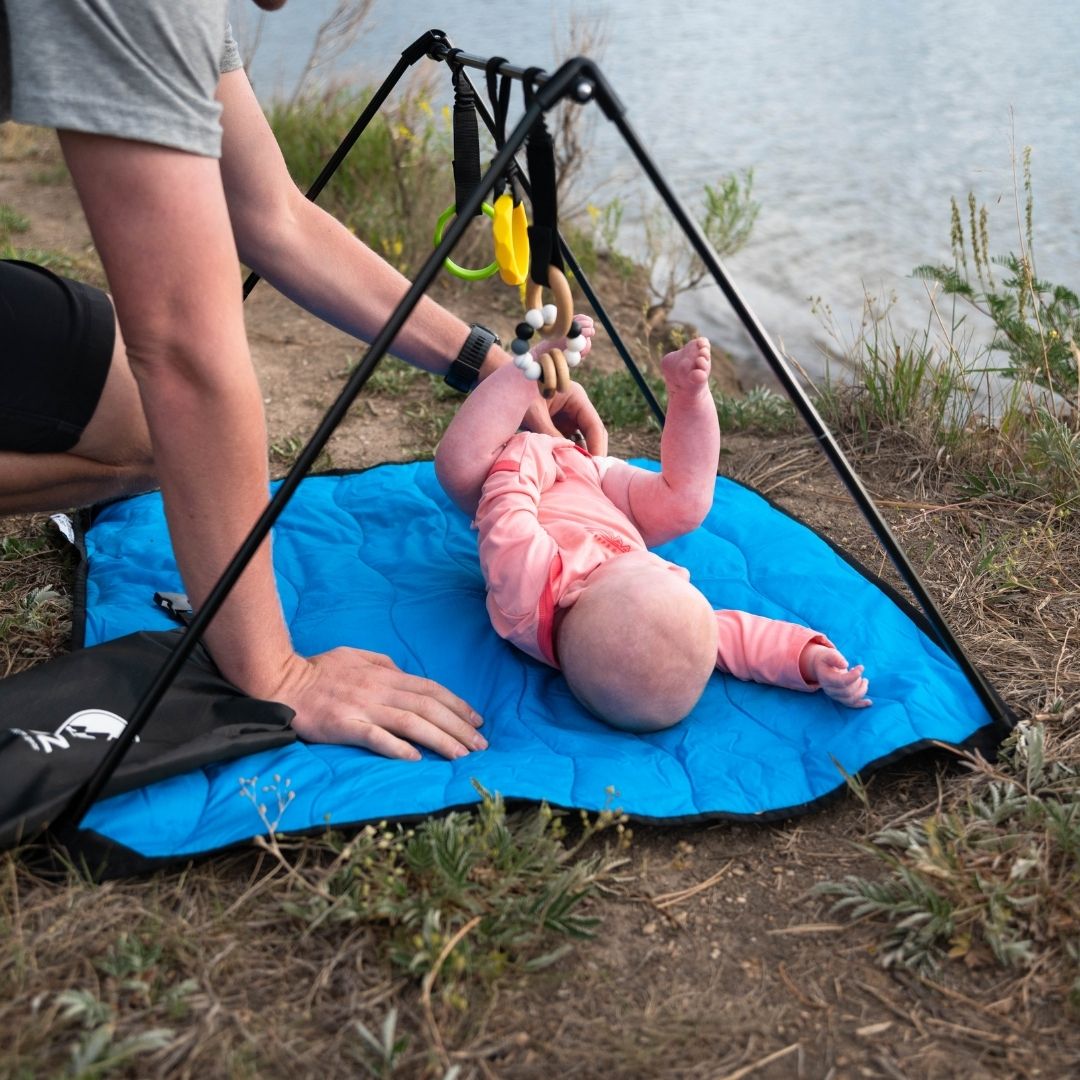 Baby on Lay and Play Adventure Mat on ground, dad's hands helping her, lake in background