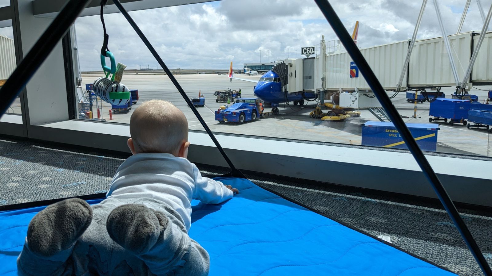 Baby lying on a blue activity gym play mat looking out of an airport window at ground operations. 