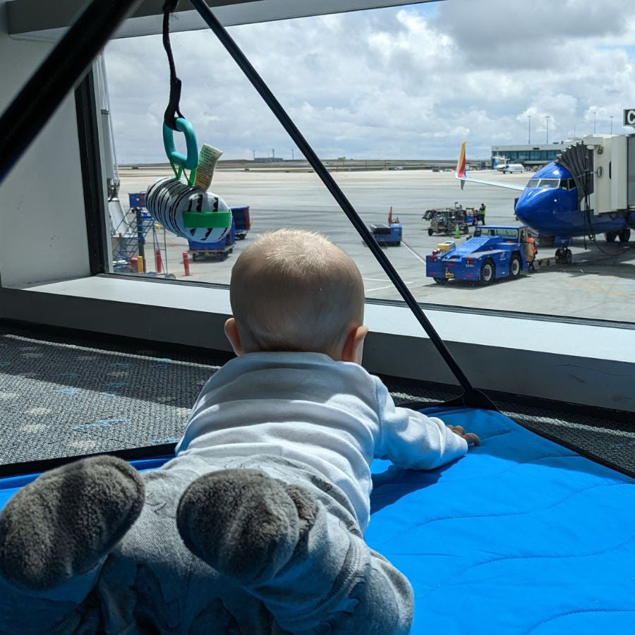 Baby lying on a blue activity gym play mat looking out of an airport window at ground operations. 