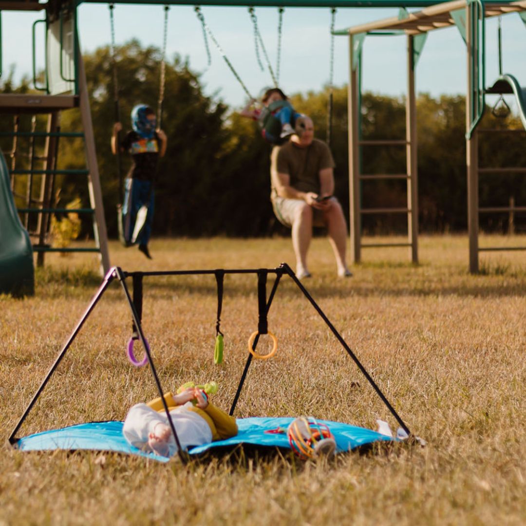  Baby laying on a baby play  mat on the grass, with people in the background and child playing on a swing set. 