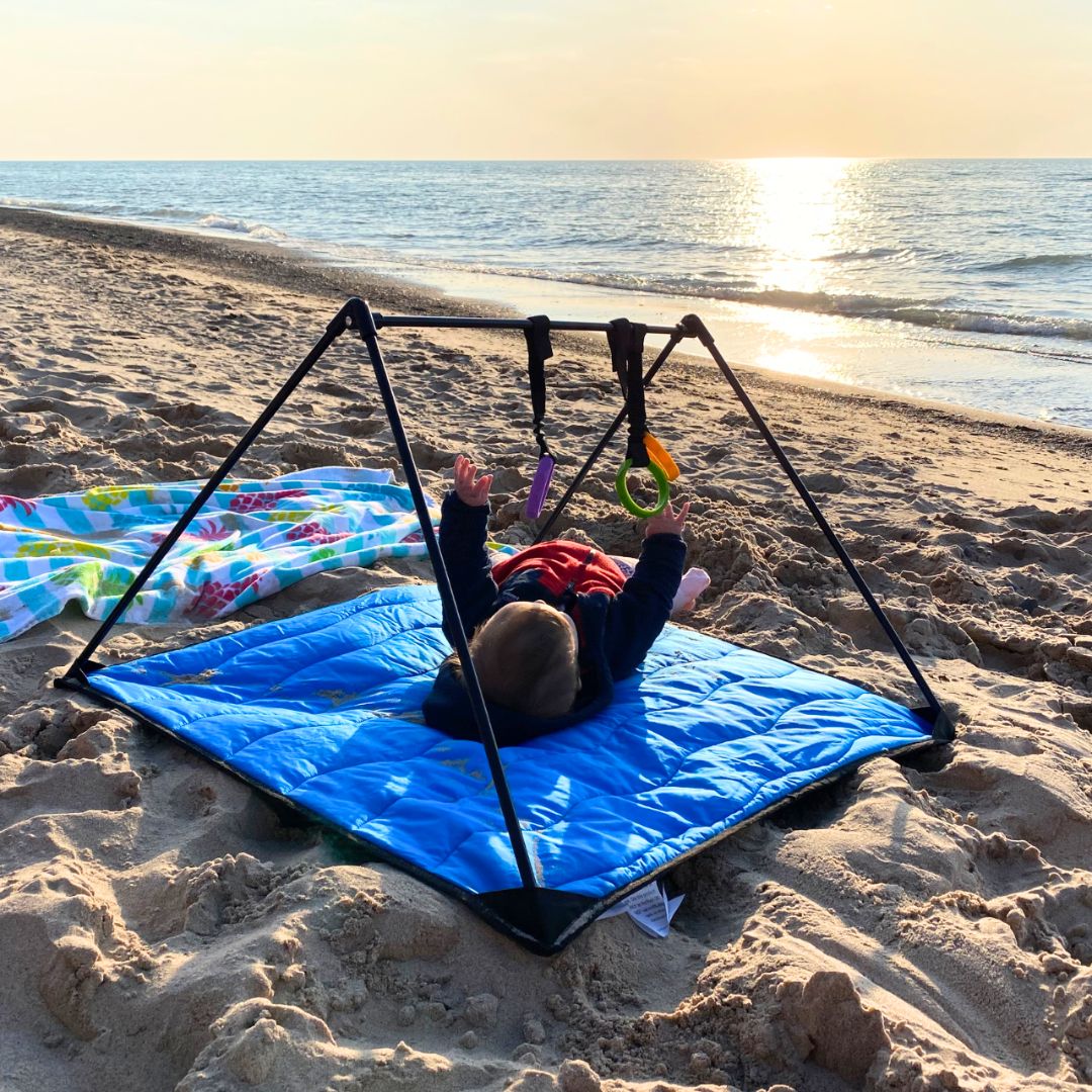 Child playing on a blue play gym mat on sand surrounded by ocean and sunset.